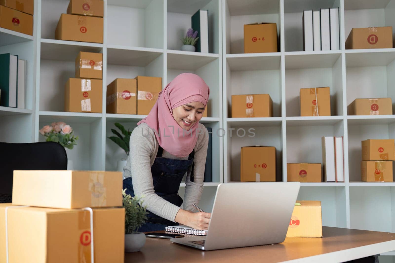 A focused Muslim woman diligently writing notes and managing her online sales in a neatly organized workspace, reflecting her entrepreneurial spirit in e-commerce.
