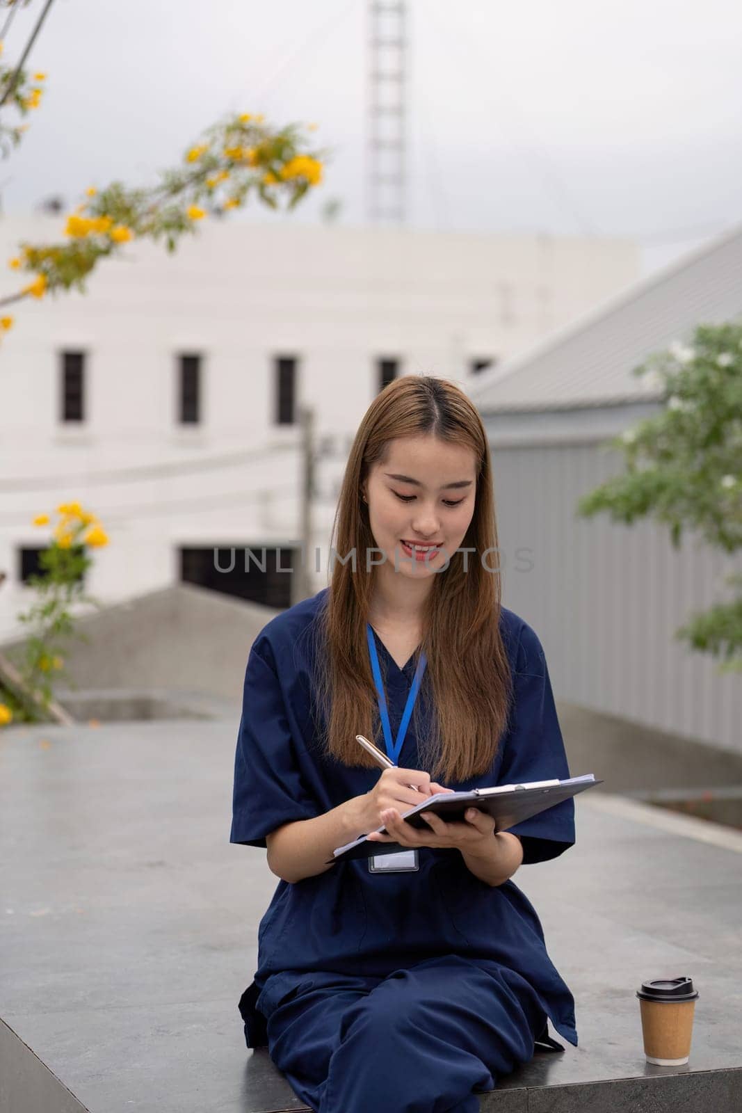 A smiling female healthcare professional in scrubs takes a break with a clipboard and coffee outdoors, emphasizing the importance of relaxation in her medical practice.