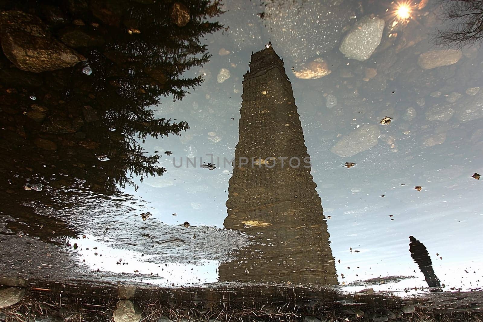 A tall stone tower stands reflected in a puddle, surrounded by scattered rocks and a person walking in the foreground.
