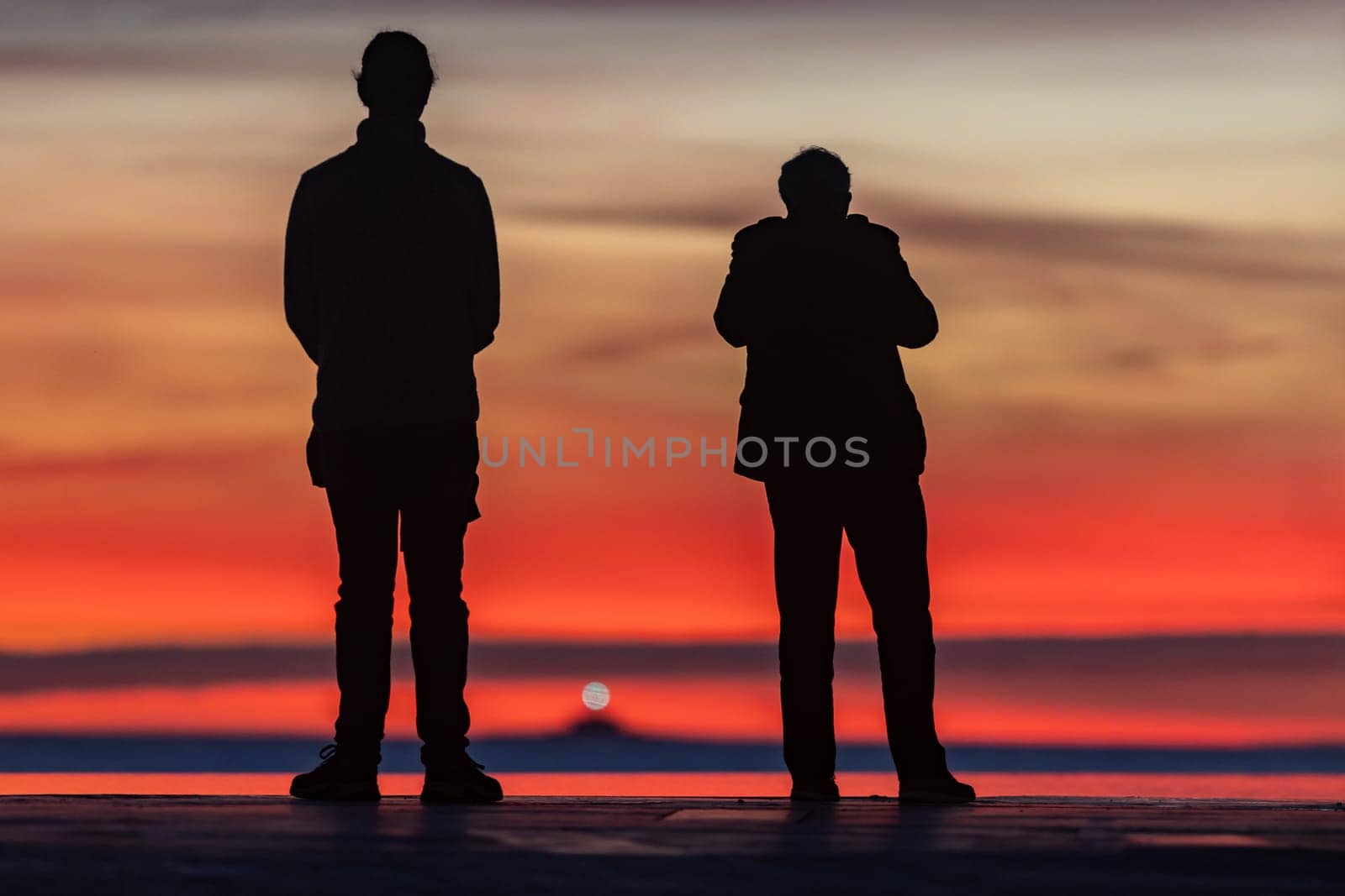 Young couple standing at the seafront and looking at bright red sunset. Mid shot