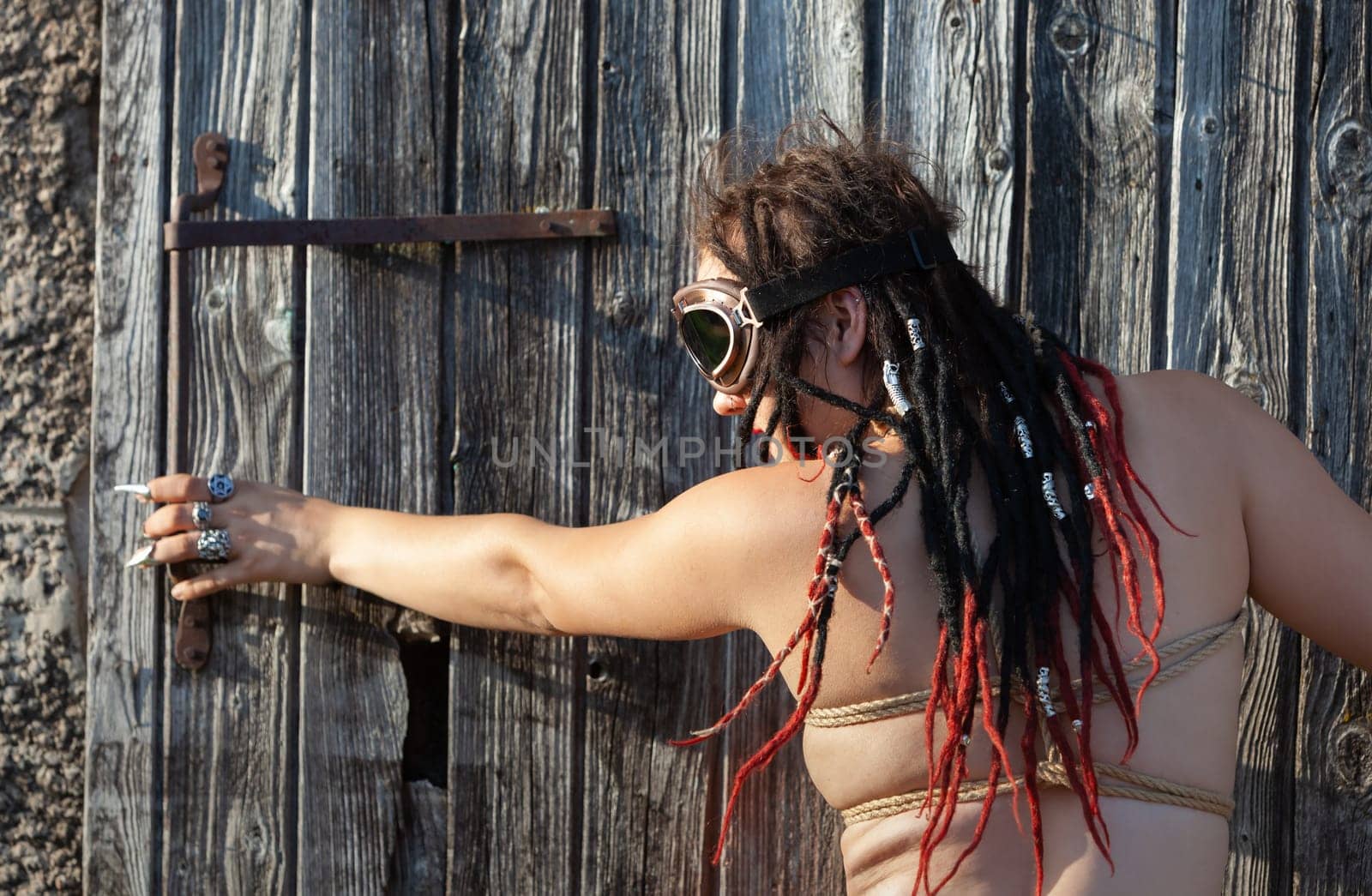 A young nude woman, roped up with dreadlocks and piercings, poses near an old wooden fence