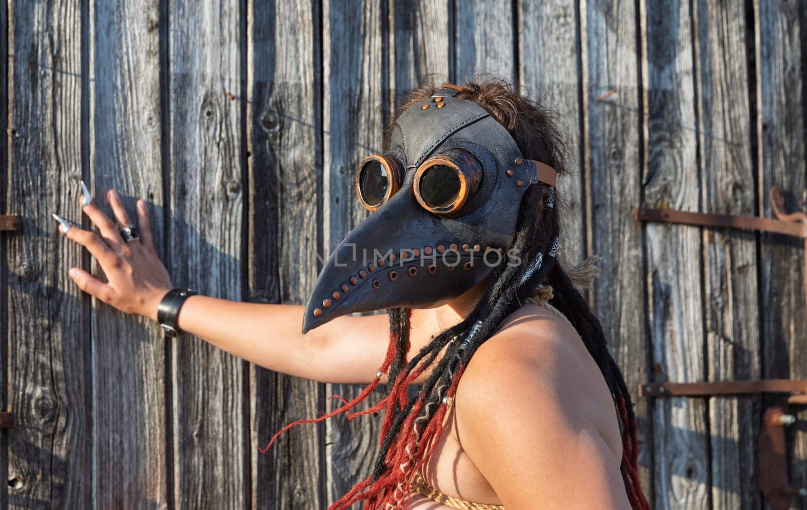 A young nude woman, roped up in a plague raven mask with dreadlocks and piercings, poses near an old wooden fence