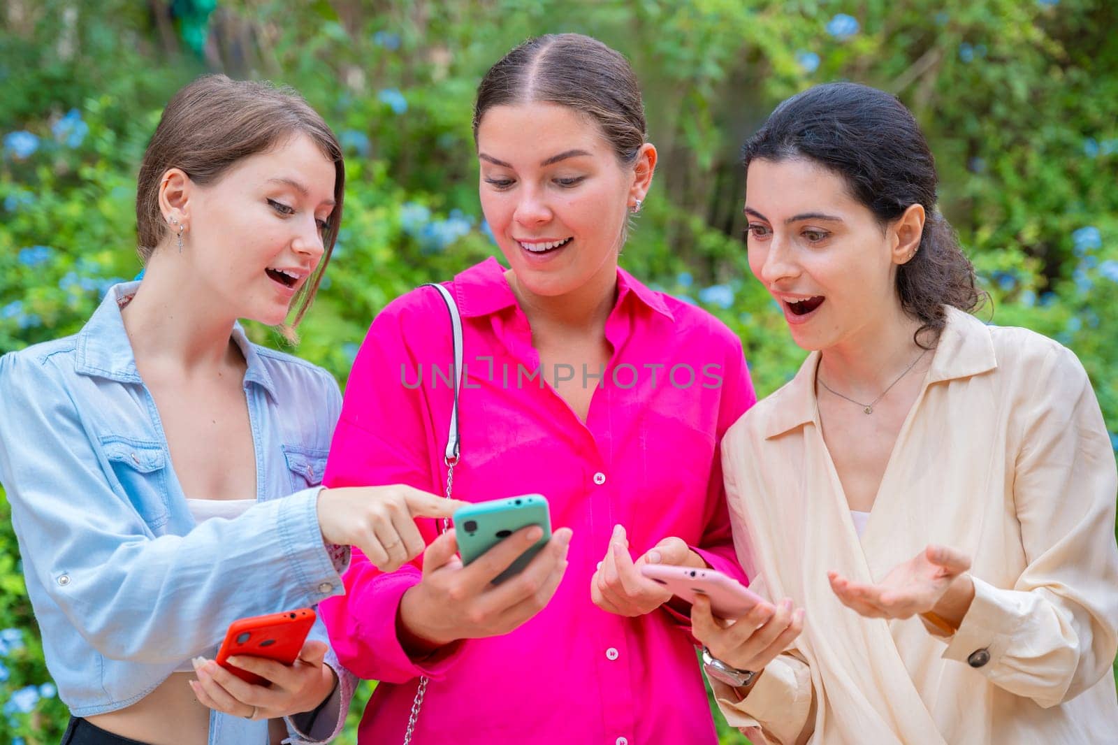 Three young women outdoors, smiling and reacting excitedly while looking at their smartphones. They appear to be sharing content or enjoying a conversation. Casual clothing and green background.