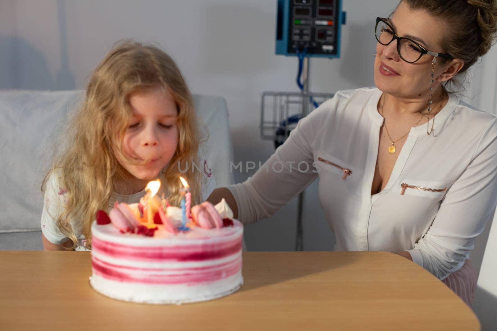 In a hospital setting, a young girl joyfully blows out the colorful candles on her birthday cake alongside her mother