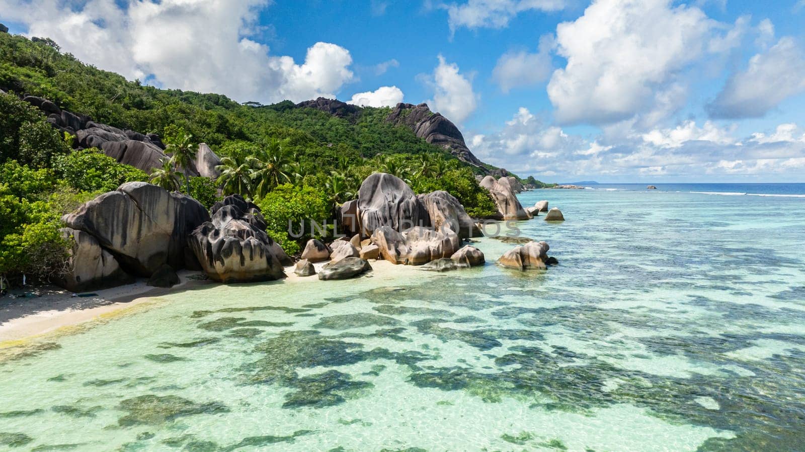Green hills rise beside clear blue waters and a sandy beach lined with rocks. Anse Source d'Argent. La Digue, Seychelles.