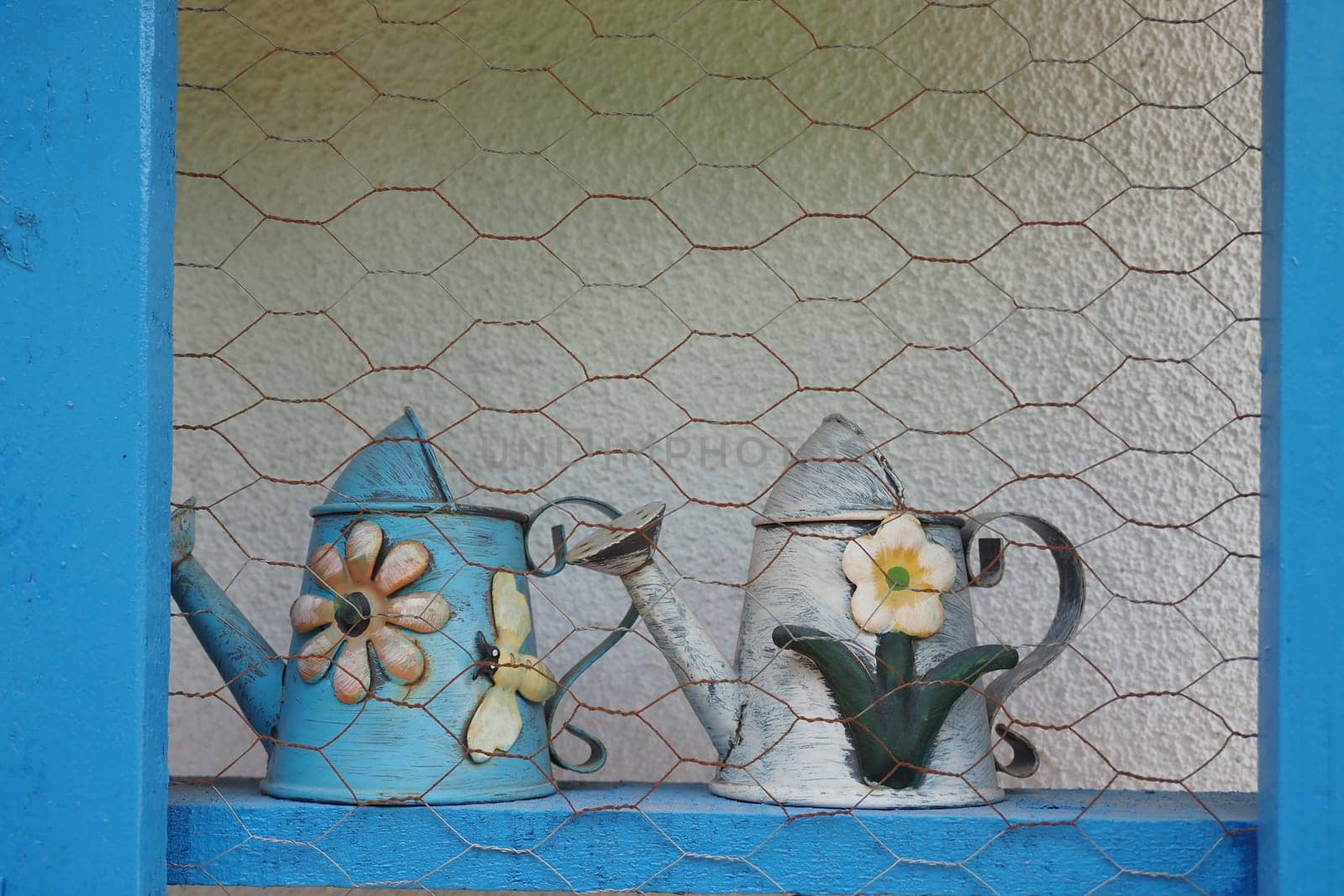 Two colorful watering cans sit on a shelf behind a mesh wire barrier.