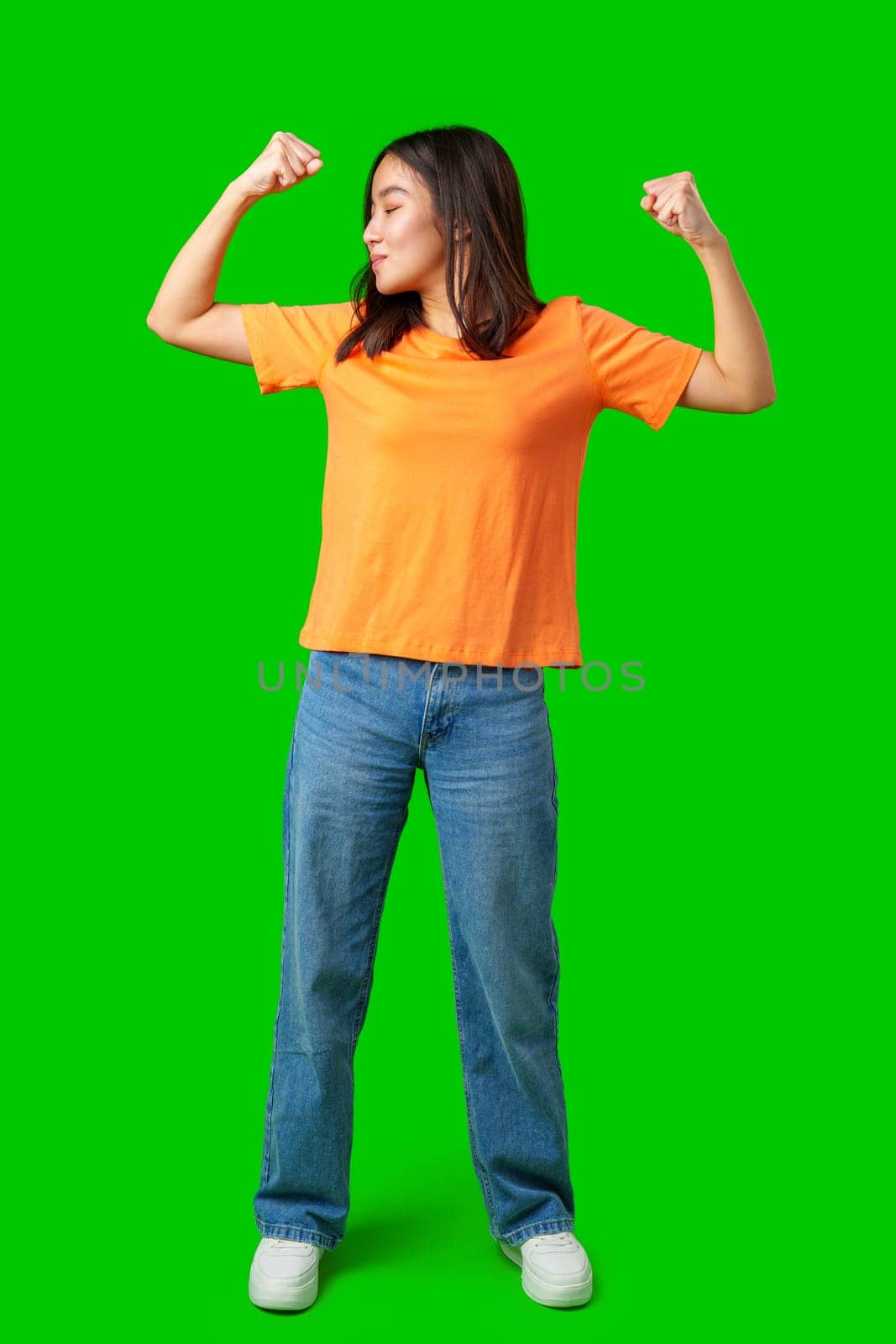 A woman stands proudly with her arms raised in a flexing pose while wearing an orange shirt and blue jeans. The bright green background adds energy to the moment.