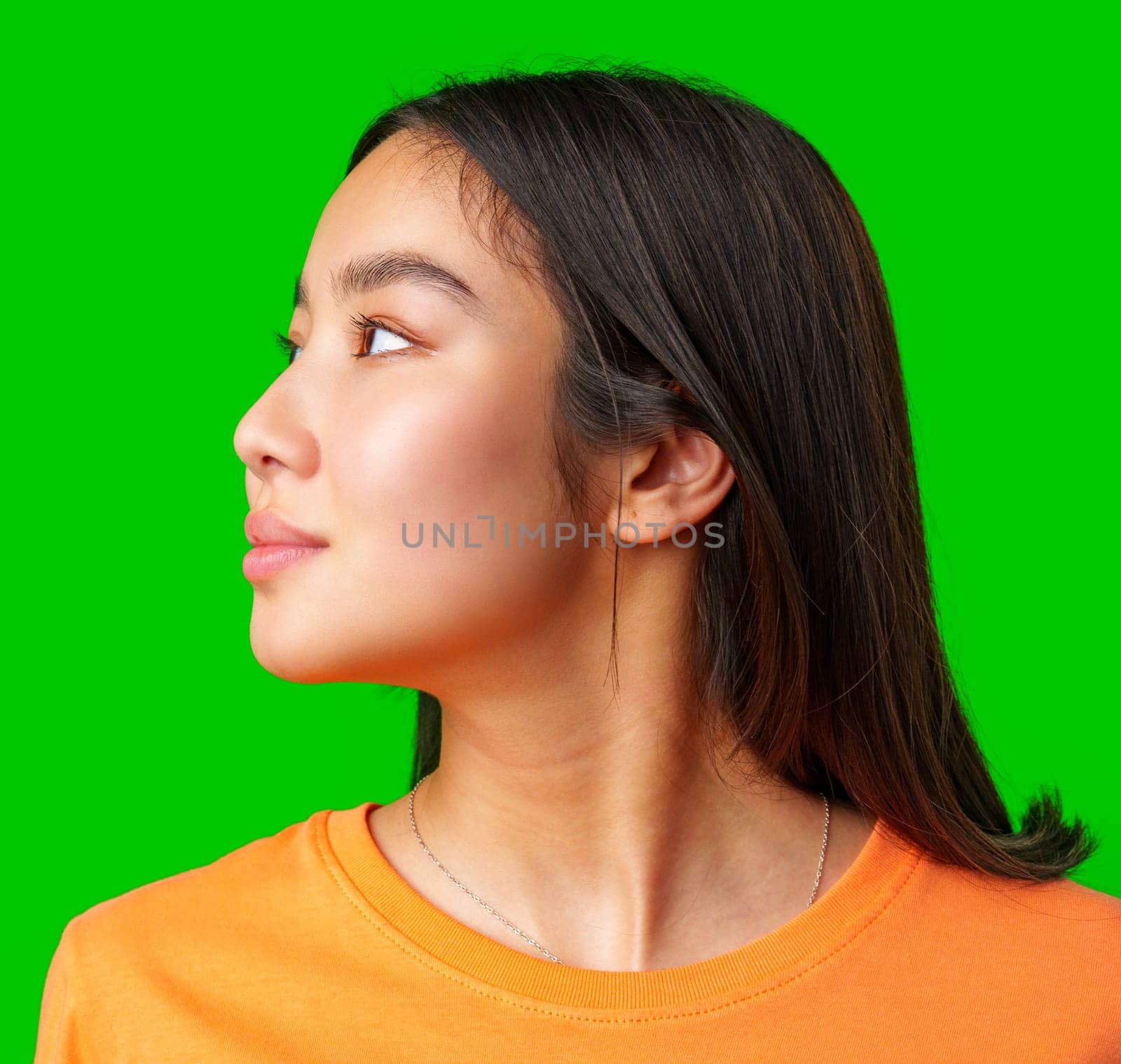 A young woman looks to the side with a thoughtful expression. She has long black hair and is wearing an orange shirt, enhancing her bright presence against a green backdrop.