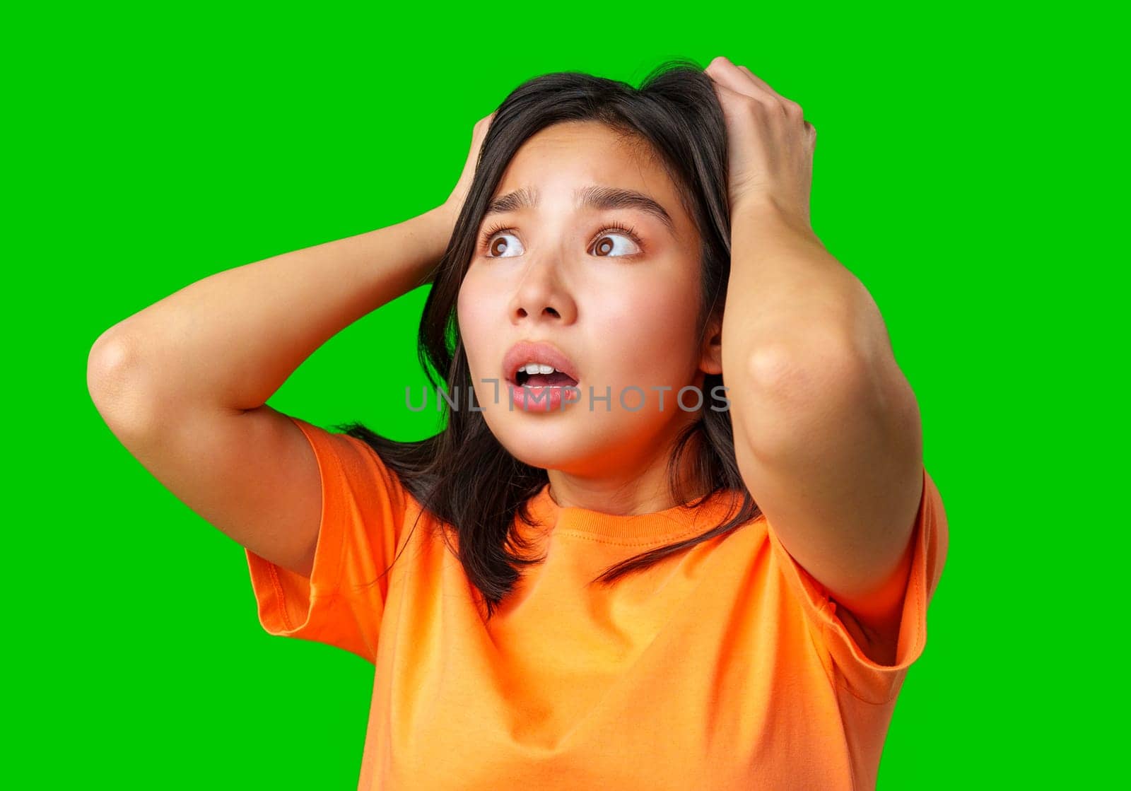 A young woman in an orange shirt expresses shock with wide eyes and hands on her head against a bright green backdrop. Her startled expression conveys disbelief or fear.