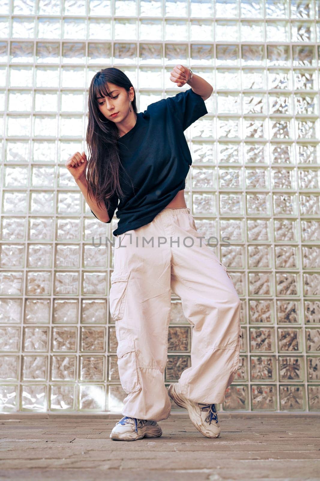 A young woman in a dark shirt and light cargo pants expresses joy through dance in front of a textured glass wall. The bright setting highlights her movements and energy.