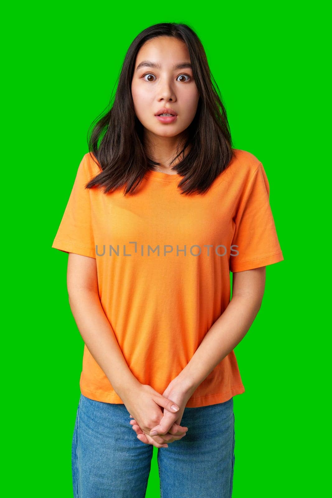 A young woman with long dark hair wears an orange shirt, displaying a surprised look while standing with her hands clasped in front of her, surrounded by a vibrant green background.