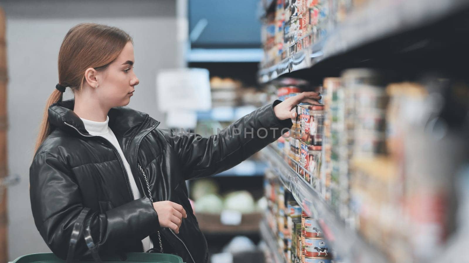 Young woman shopping groceries, reaching for a can on a supermarket shelf, making purchasing decisions in a retail environment