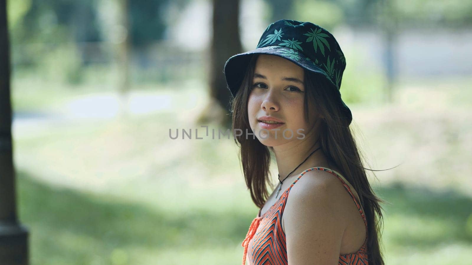 Portrait of a teenage girl wearing a bucket hat featuring a marijuana leaf design, posing in a park during a sunny summer day
