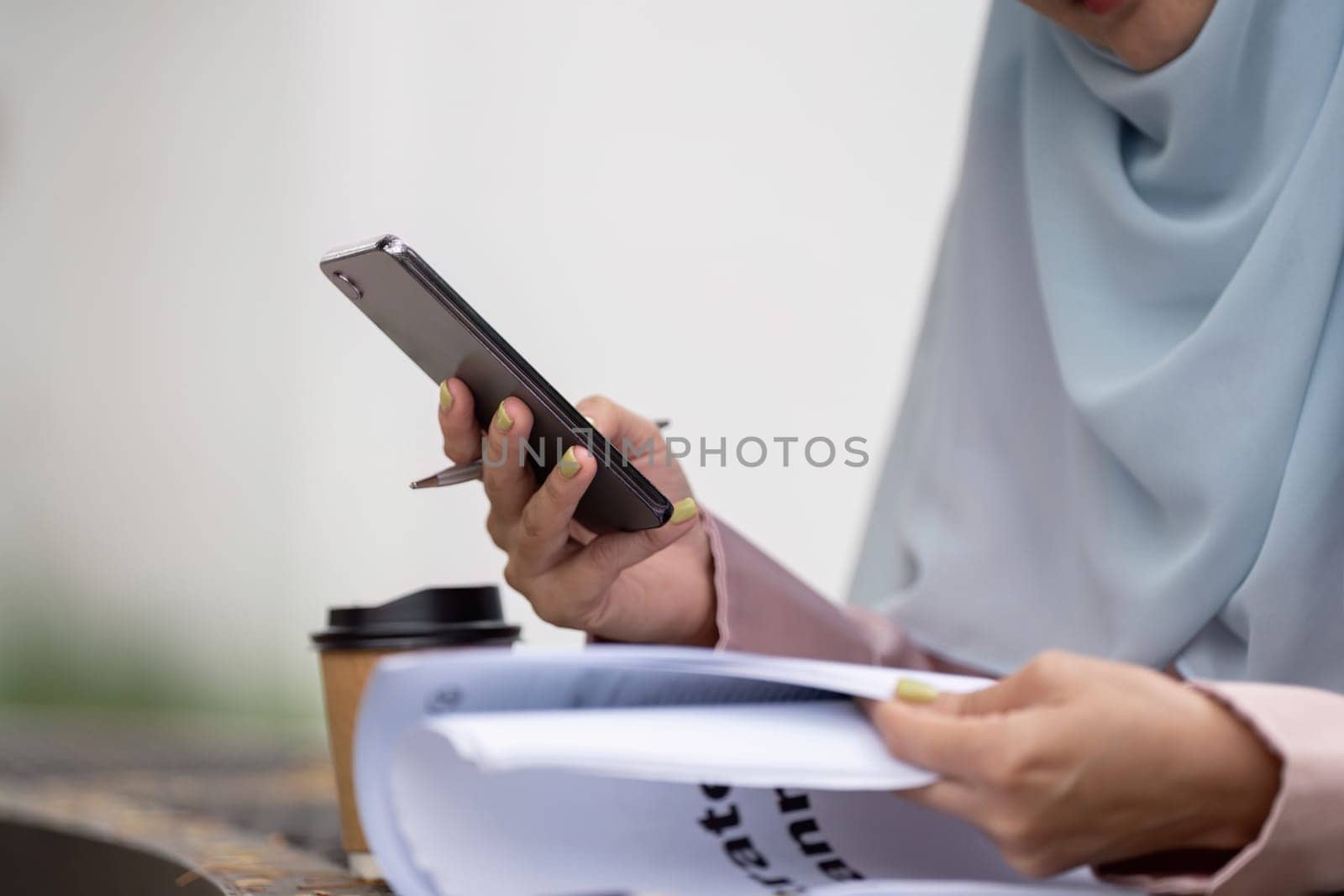 A Muslim businesswoman in hijab checks her smartphone while reviewing documents and holding a coffee cup, reflecting multitasking in a professional environment.