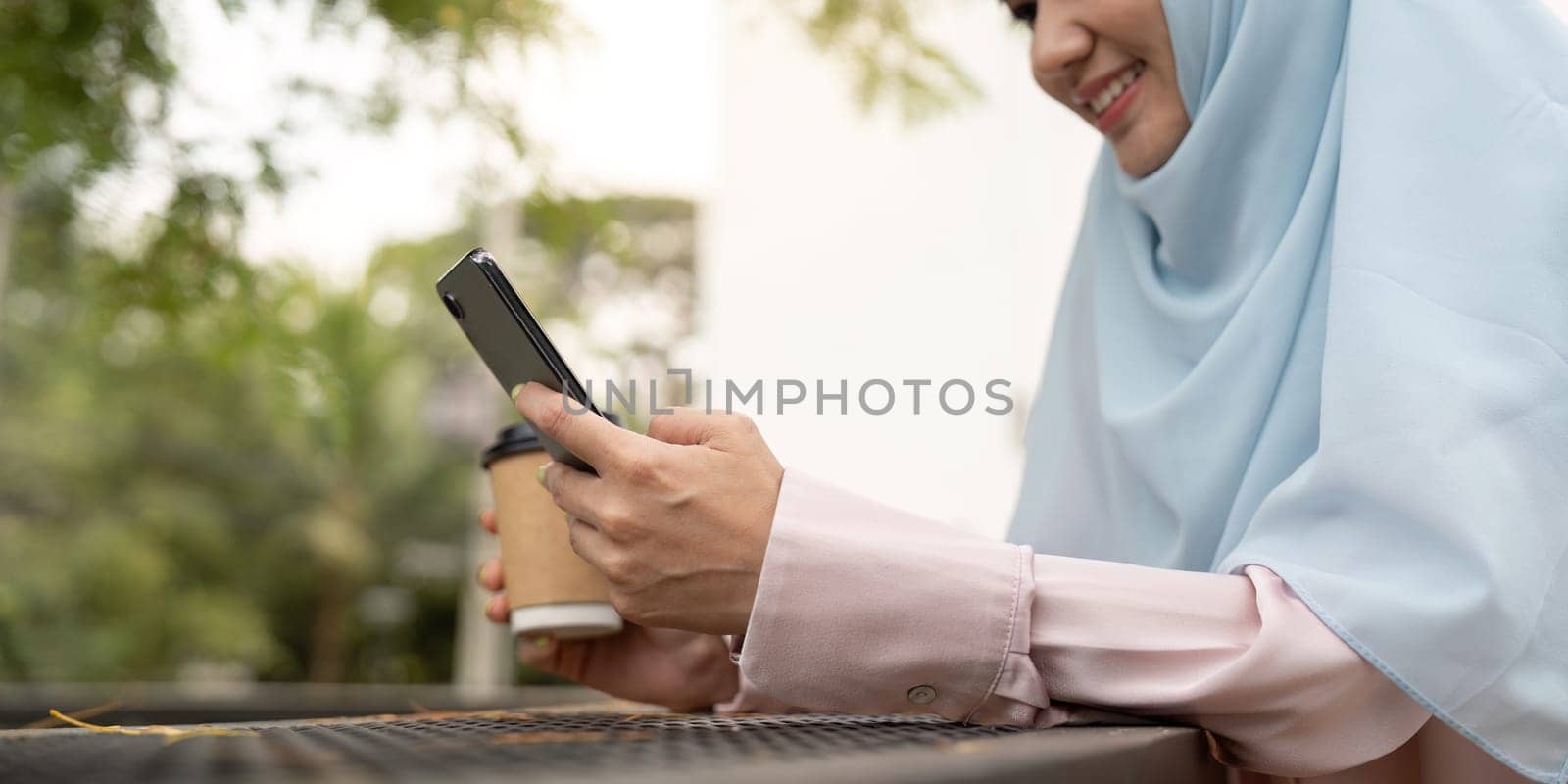 A Muslim businesswoman in hijab smiles as she uses her smartphone and holds a coffee cup, enjoying a relaxed moment in an outdoor setting.