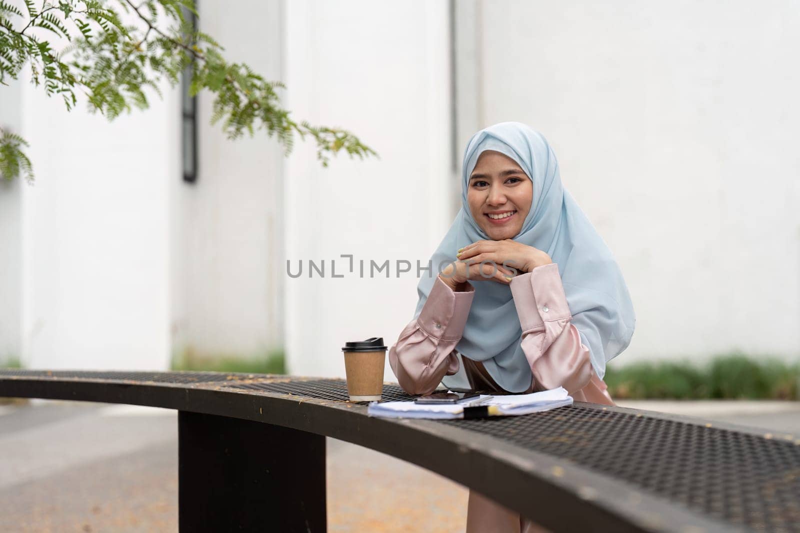 A Muslim businesswoman in a hijab smiles while resting her hands on a table with a coffee cup and documents, enjoying her time in an outdoor professional setting.