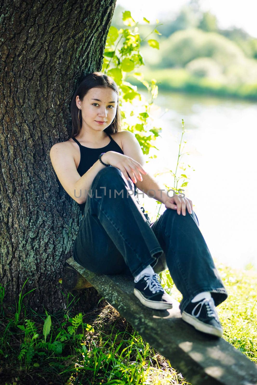 Serene moment of peace as a young woman enjoys the tranquility of nature, sitting on a wooden plank beneath a large tree by a river