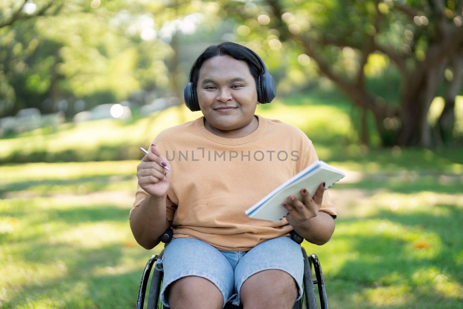 A diverse young man in a wheelchair, smiling and holding a sketchpad with a pencil, wearing headphones in a vibrant park setting.