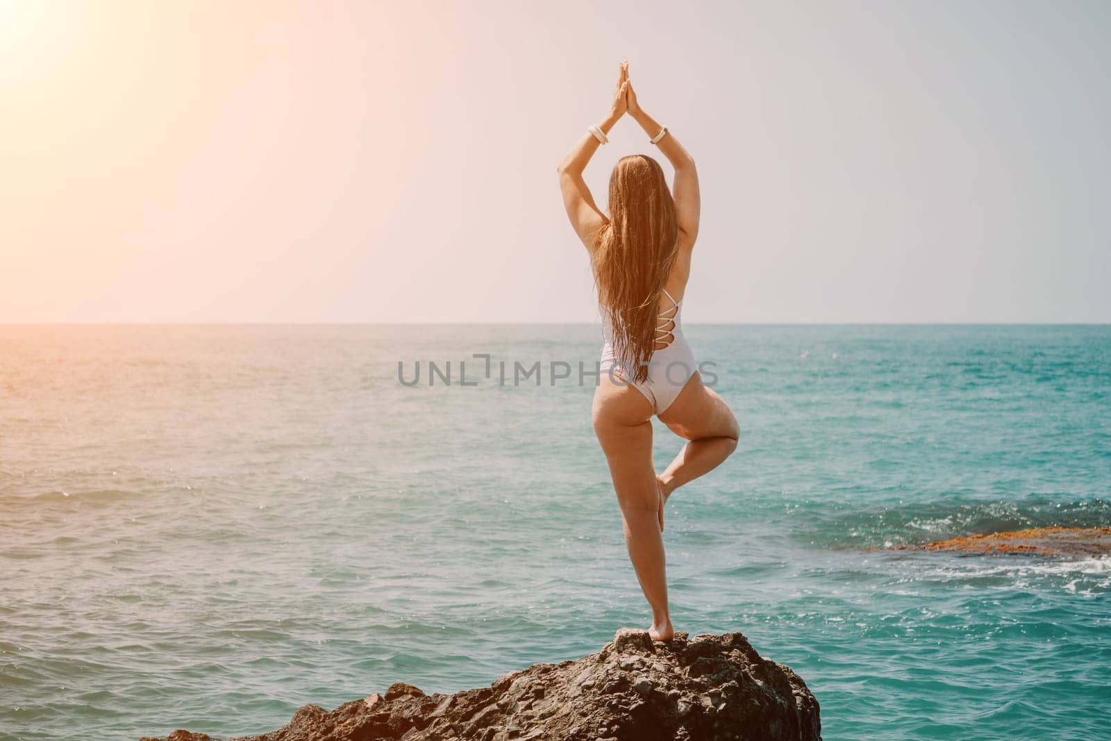 Woman sea yoga. Back view of free calm happy satisfied woman with long hair standing on top rock with yoga position against of sky by the sea. Healthy lifestyle outdoors in nature, fitness concept. by panophotograph