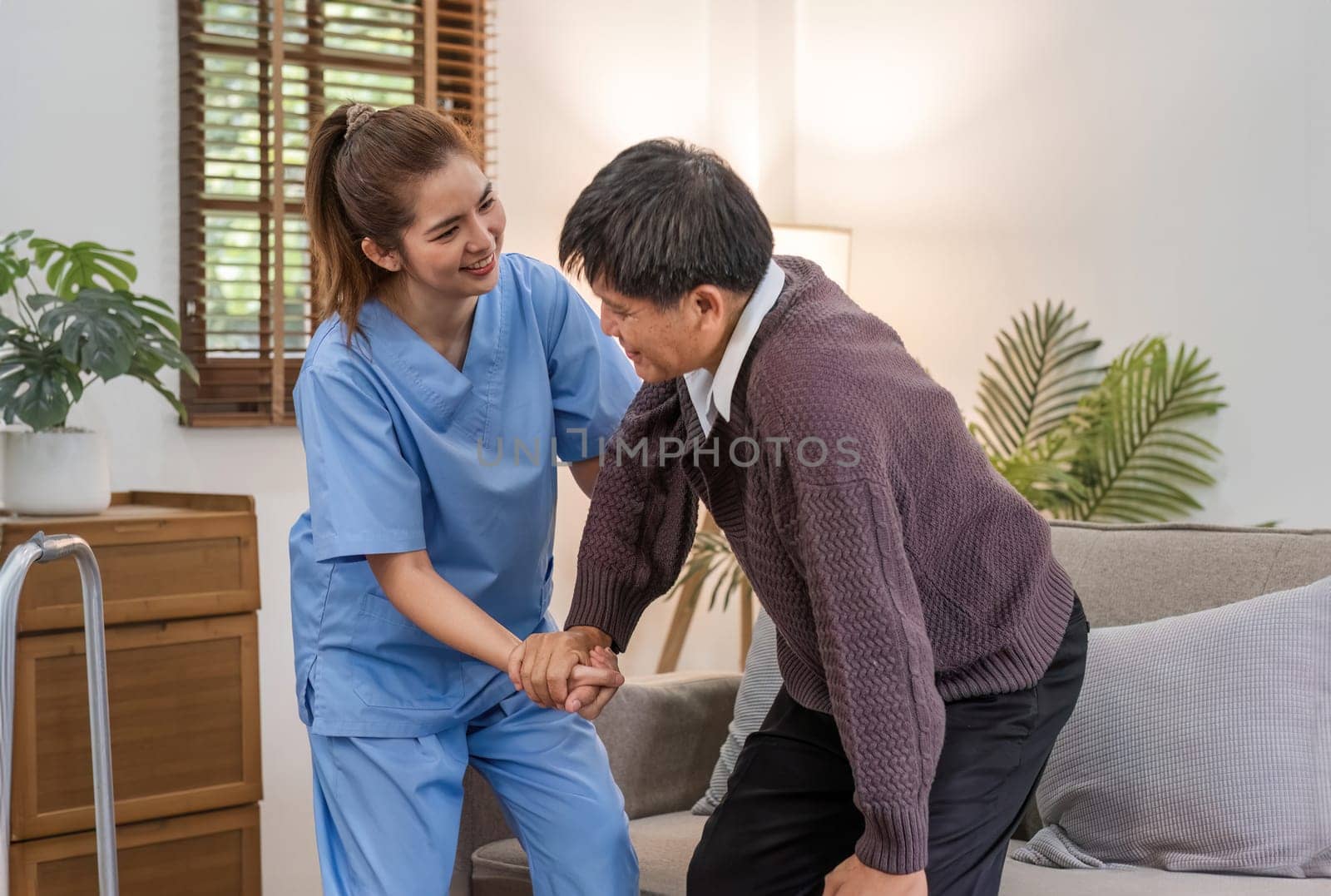 A caring young Asian nurse helps an elderly person walk in the house. An elderly disabled man tries to walk with the help of a female caregiver. by wichayada