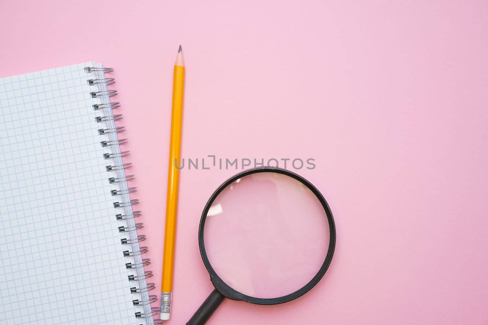 Magnifying glass, pencils and a blank white notepad on a spring on a pink background. Back to school concept. Copy space for text