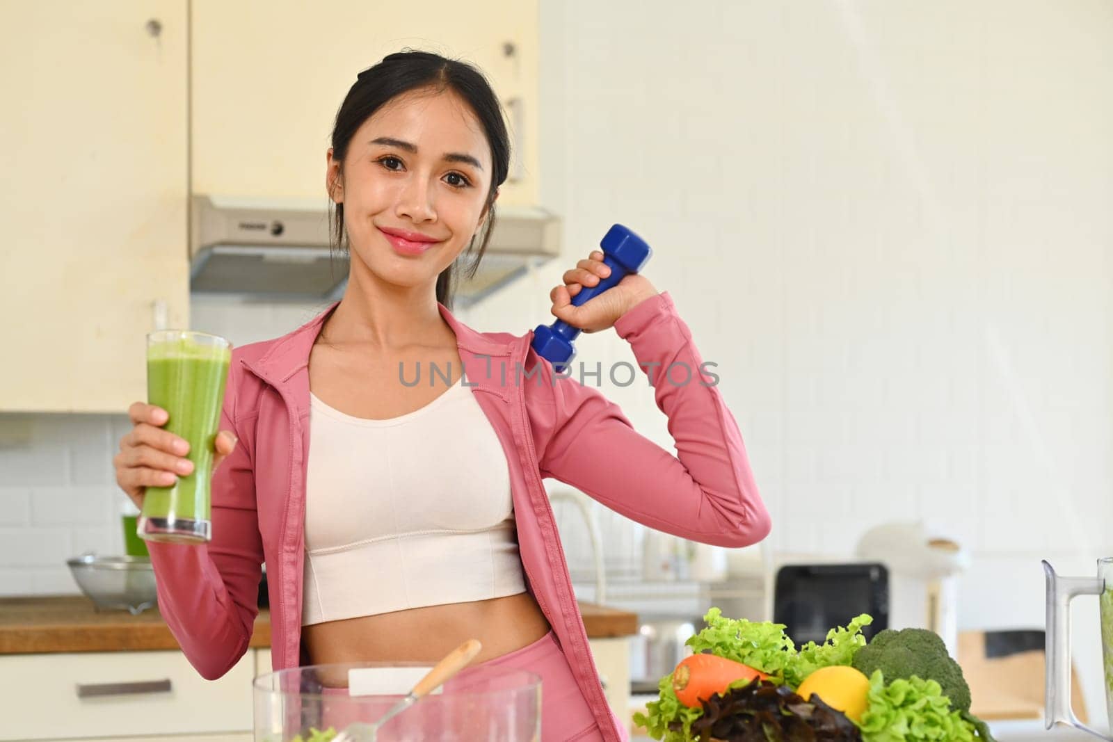 Young fitness woman holding glass of fresh green vegetable juice and dumbbell in kitchen. Healthy lifestyle concept.