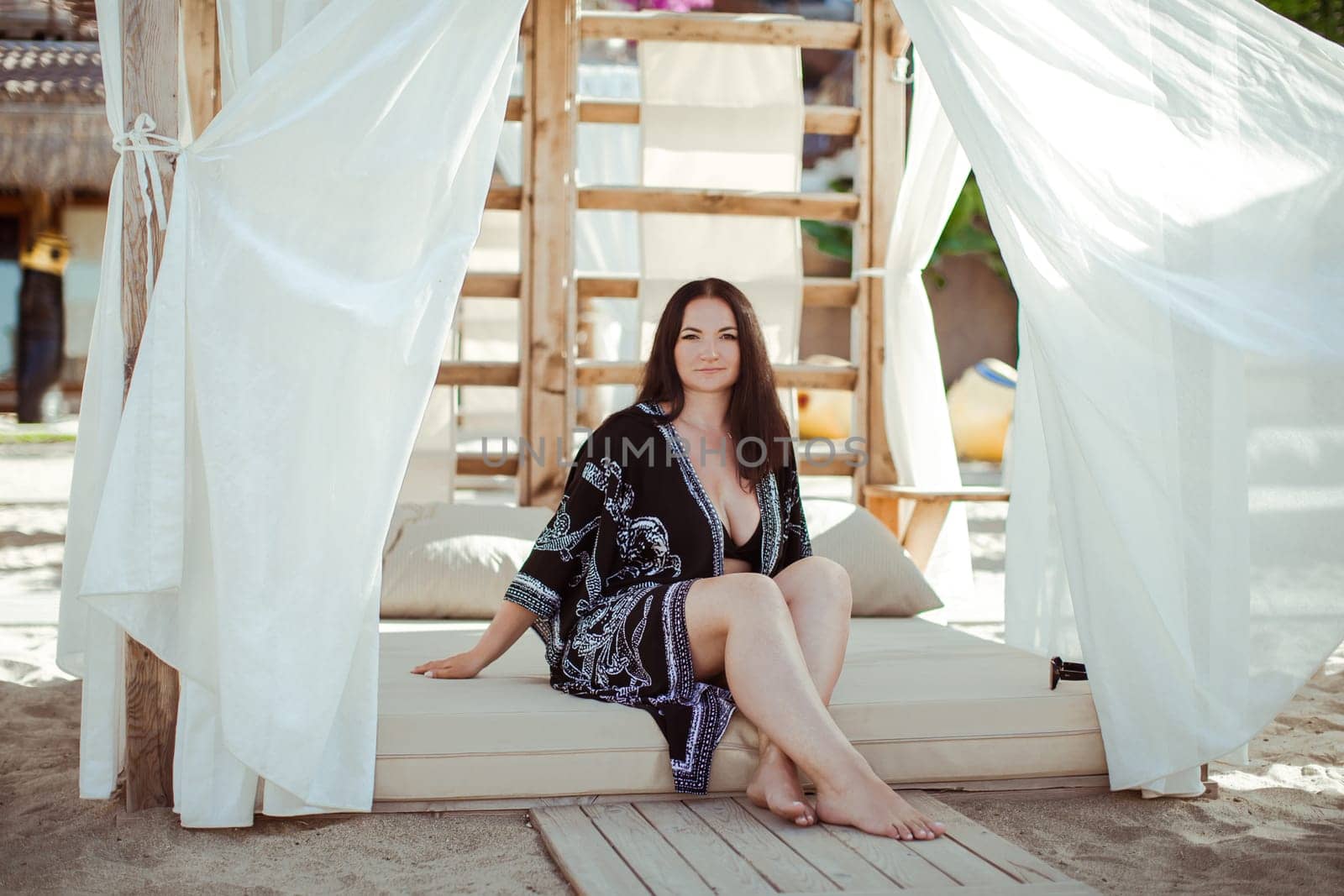 A brunette girl in a black cape with an oriental ornament is sitting in a beach tent