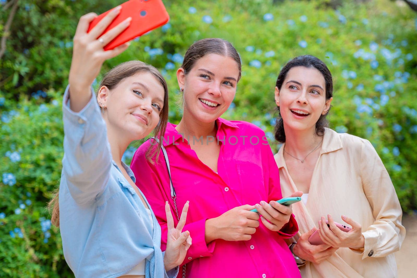 Three smiling women posing for a selfie in a lush green park. They embrace each other, expressing happiness and friendship. The vibrant colors and natural background create a joyful atmosphere.