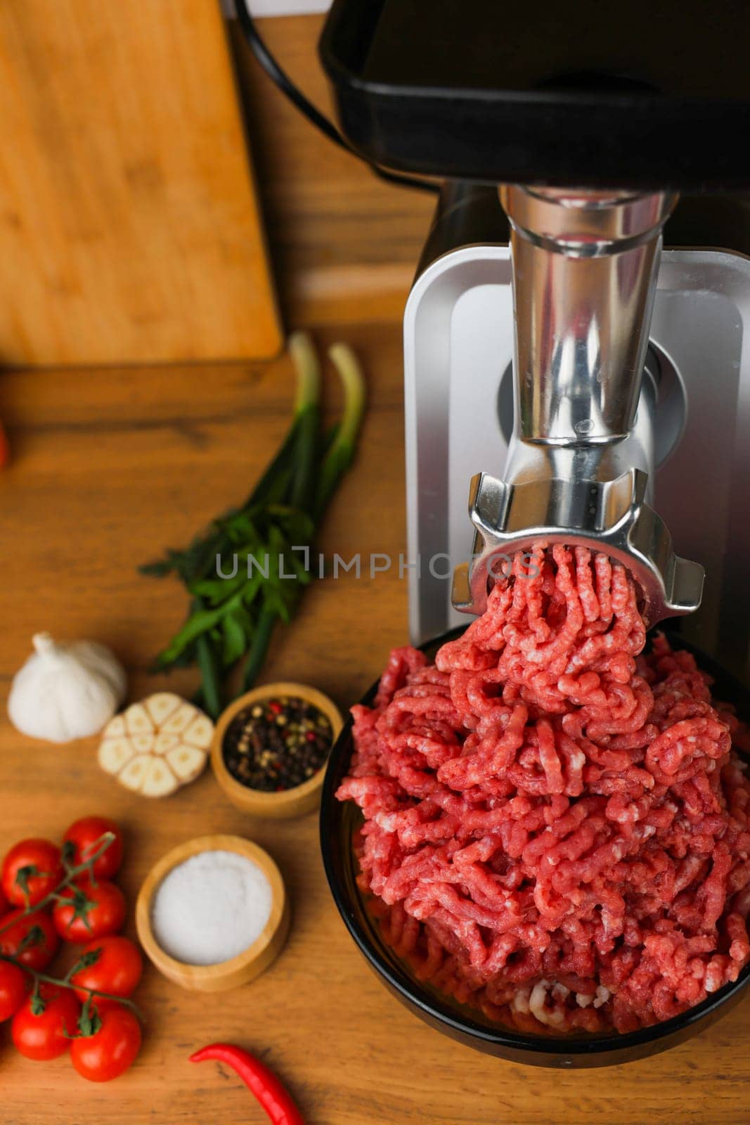 Vertical photo view from above, on a wooden background, on the right there is a metal meat grinder and a black dish into which red minced meat comes out of a meat grinder, spices, herbs and vegetables are nearby. High quality photo