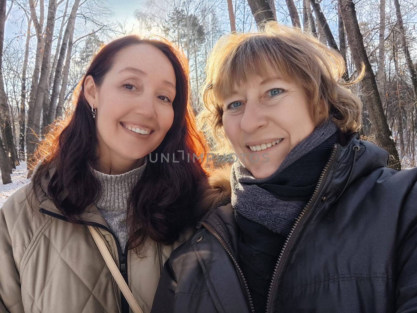 Two friends smile for a selfie while walking through a snowy forest, enjoying each other's company on a bright winter day.