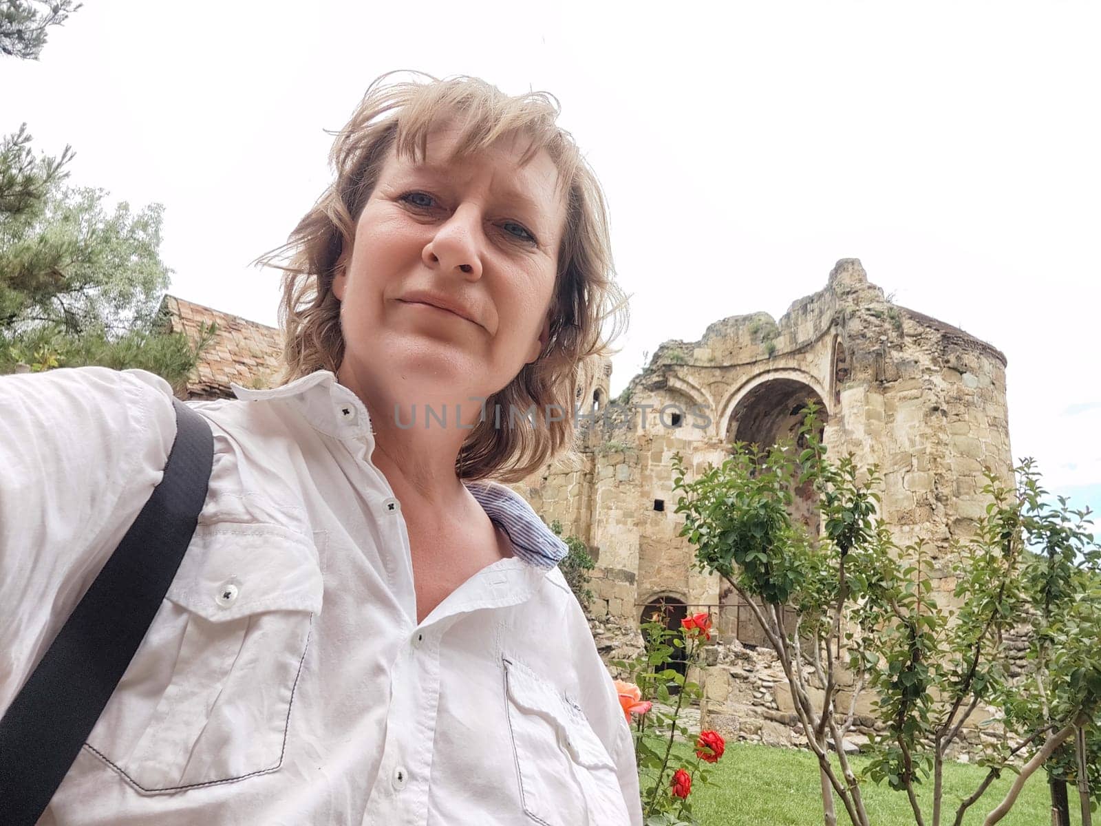 A female travel blogger captures a selfie at a historical site surrounded by lush greenery and vibrant flowers on her journey.