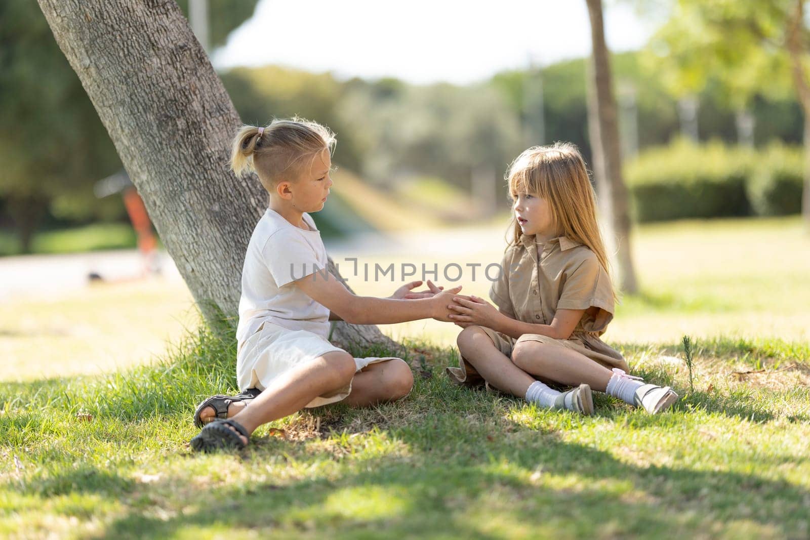 Two children sitting on grass, talking under a tree in a sunny park