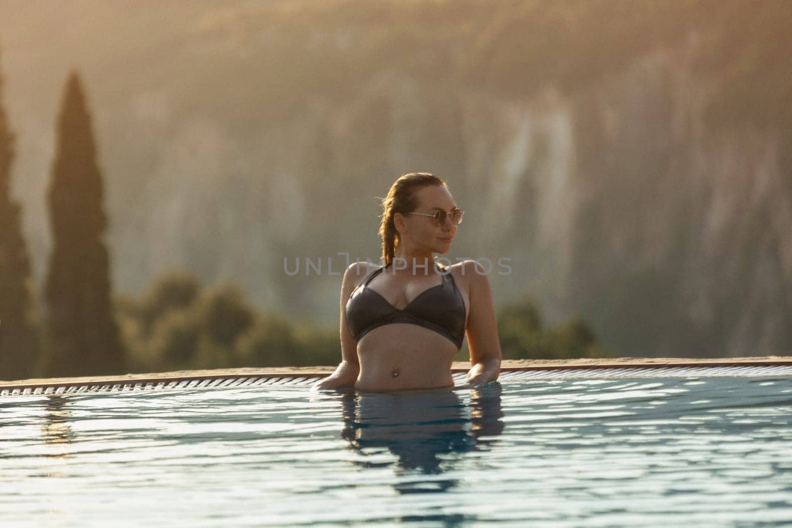 Slim woman in swimsuit walking by the edge of luxury infinity pool with jungle view .