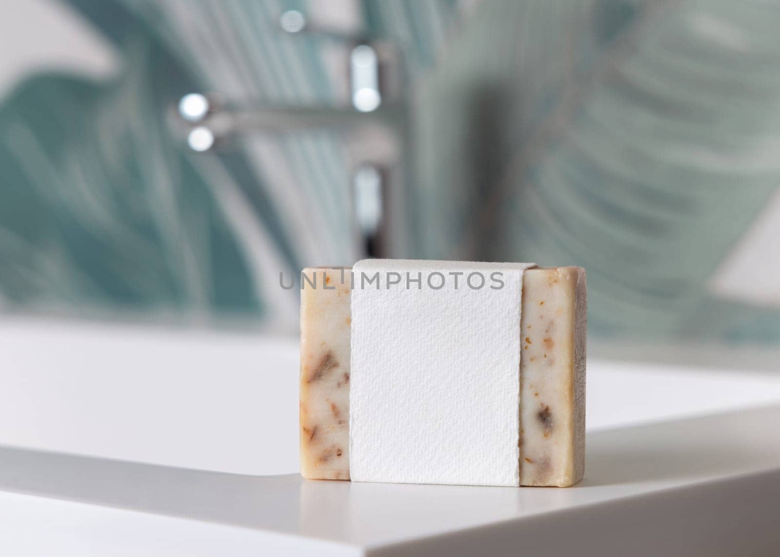 Beige soap bar with blank label on basin near faucet in green tropical bathroom close up, packaging mockup with copy space. Natural plant unbranded product for daily hygiene
