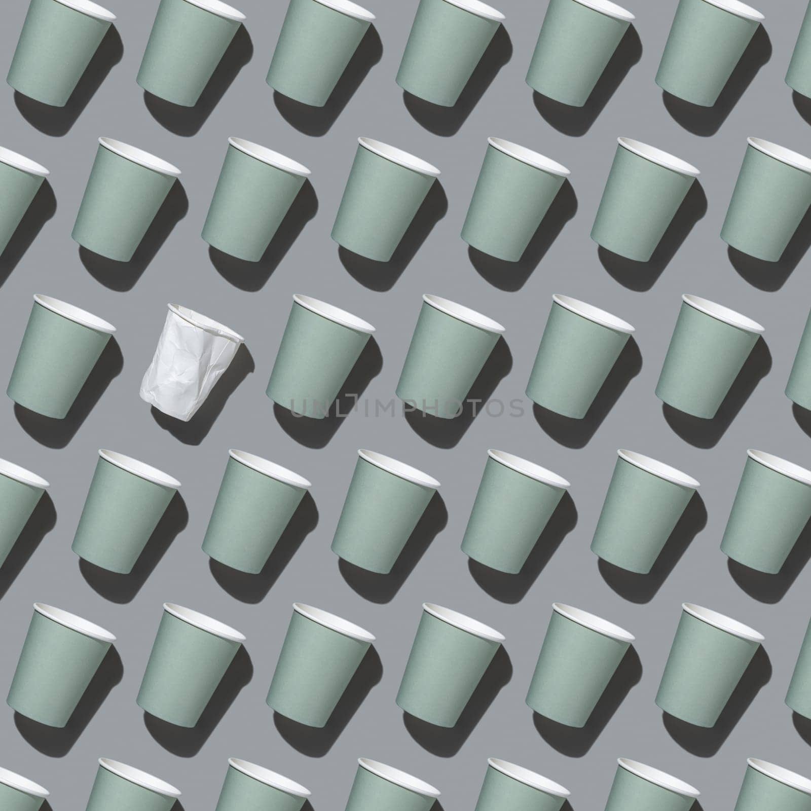 Disposable new light green paper cups and crumpled used white one in a line on gray top view, pattern, hard shadow, mockup with copy space. Empty cardboard coffee cups flat lay. Consumerism concept