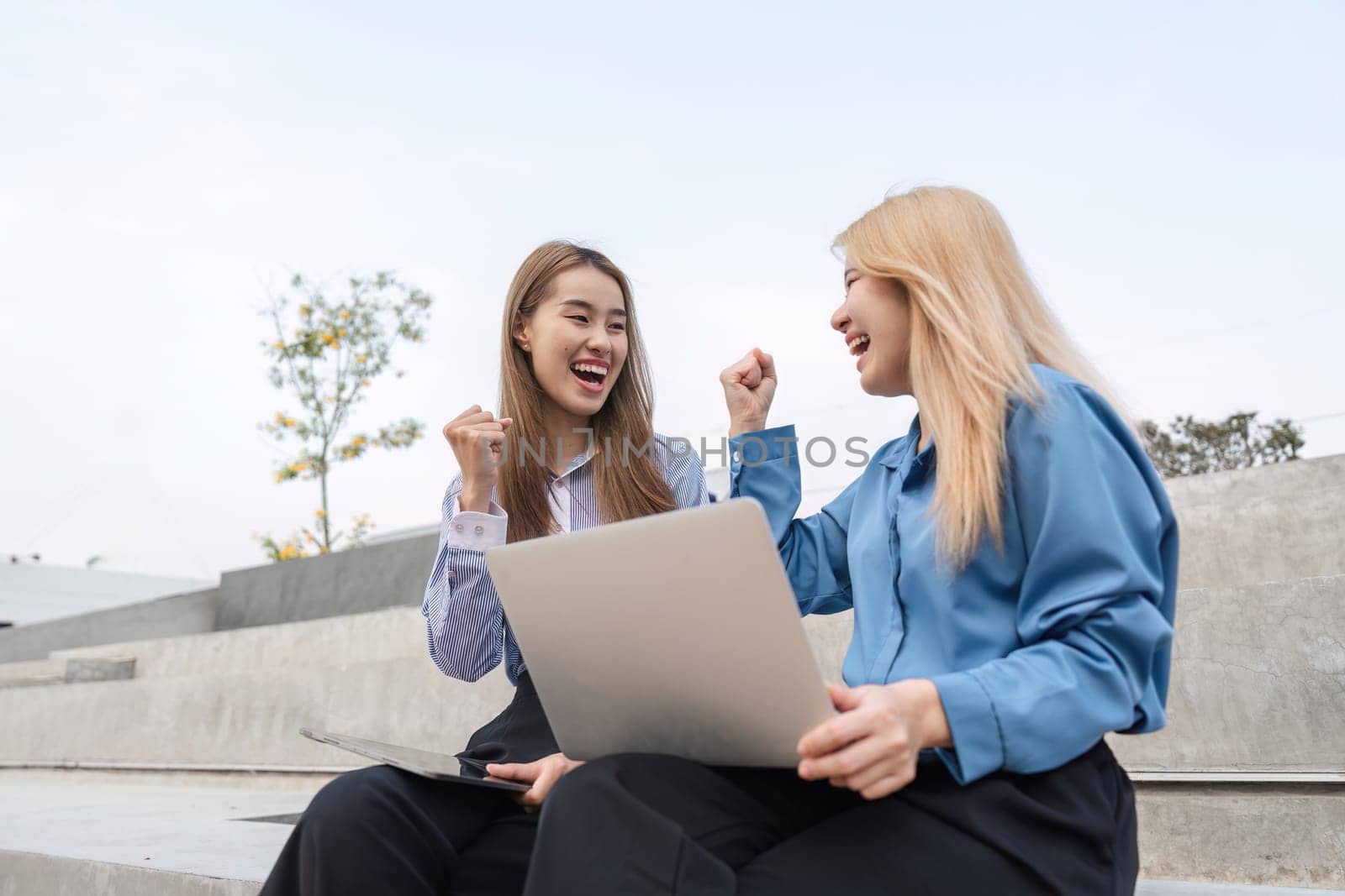 Two businesswomen outdoors joyfully celebrating their success, raising their fists in excitement while sitting with laptops on outdoor steps.
