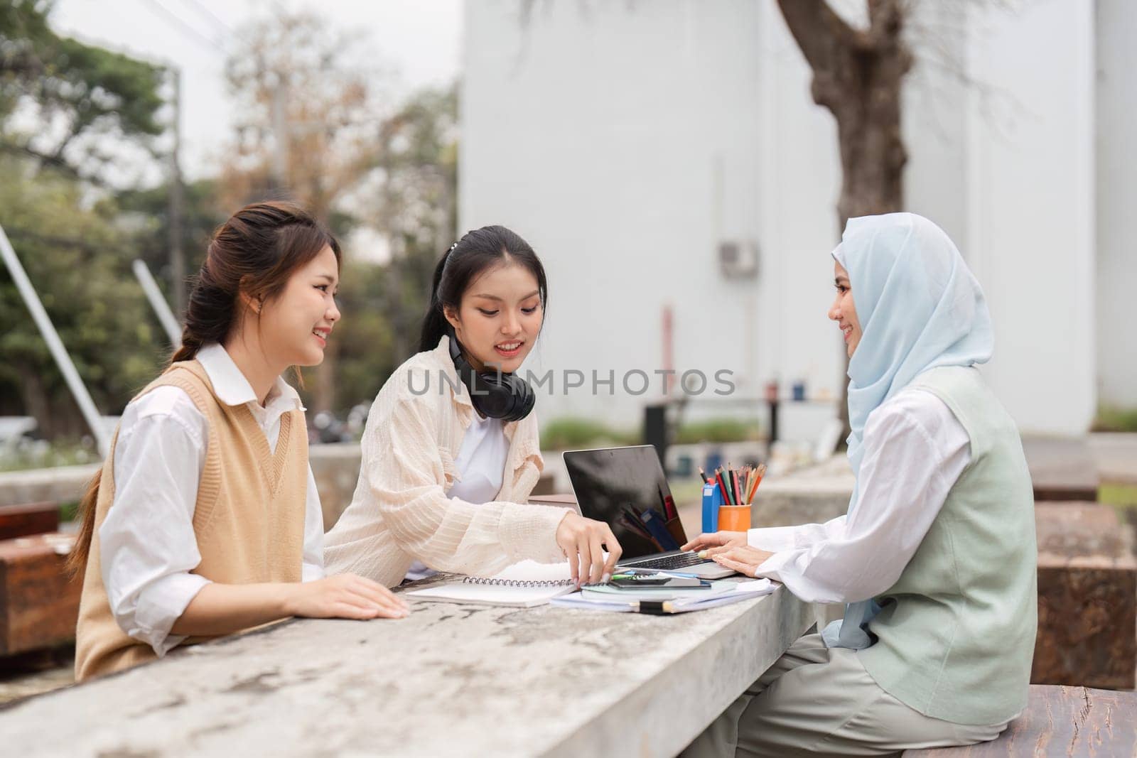A diverse group of three women, including one in a hijab, collaborating on projects outdoors, fostering teamwork and sharing ideas in an academic environment.
