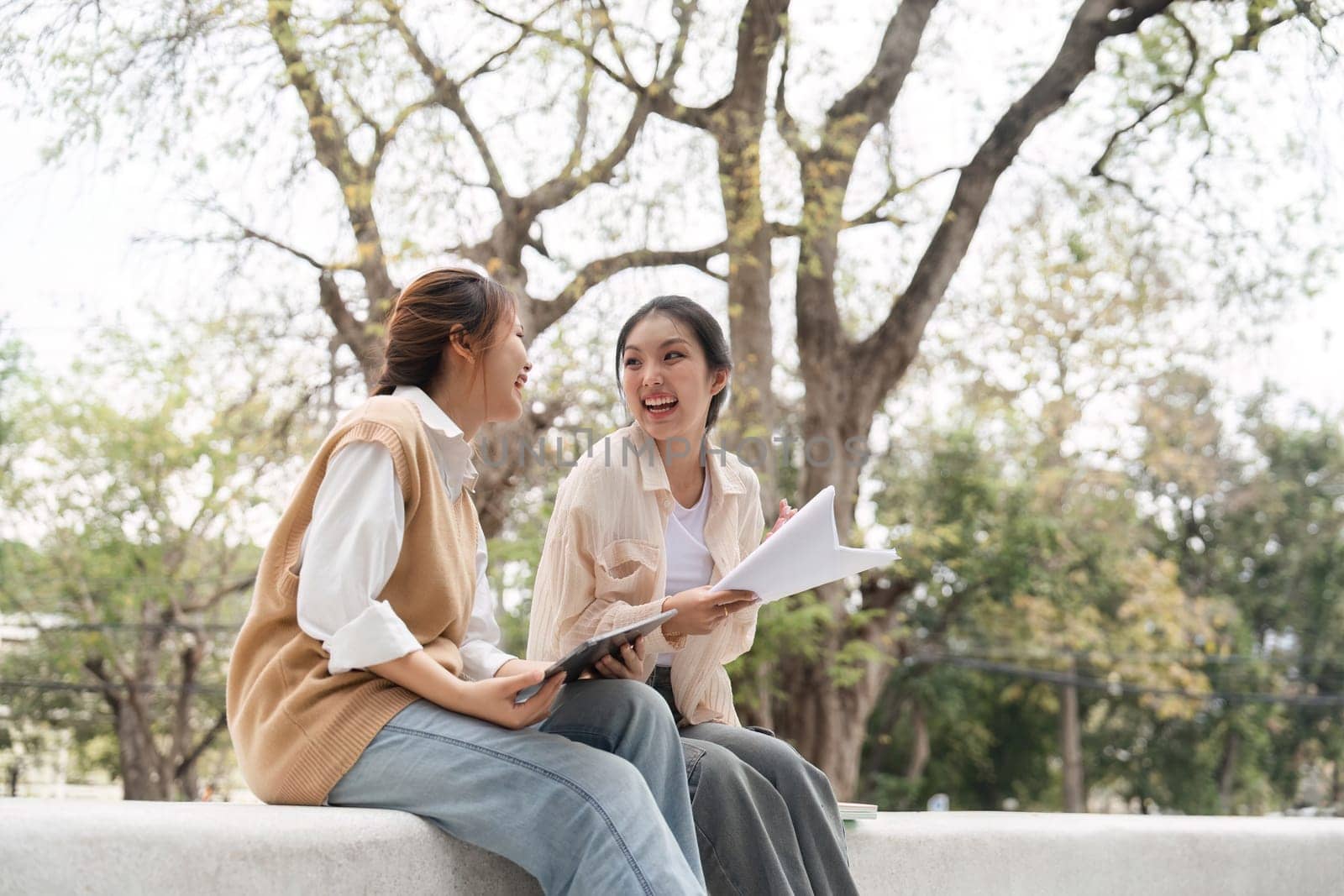 Diverse female students sharing a joyful moment while studying together, using notes and a tablet in a peaceful outdoor setting on their university campus.
