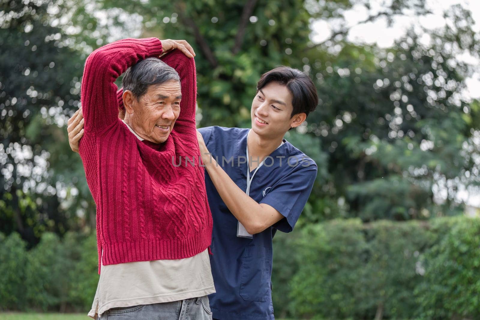 In a vibrant garden, a young caregiver helps an elderly man with his stretching exercise, promoting health and strength in a warm and caring environment.