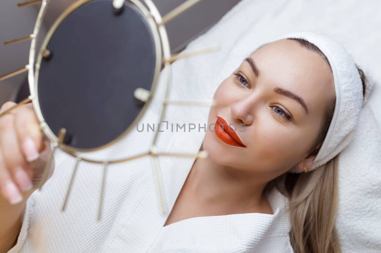 Permanent lip tattooing. A sexy girl with beautiful lips looks into the mirrors lying on the couch at the cosmetologist.