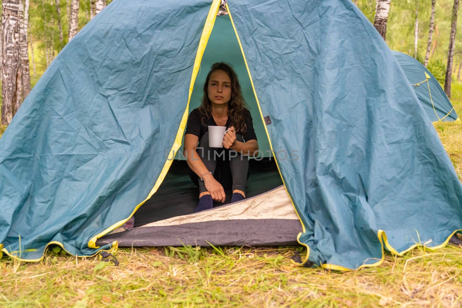 Woman relaxing inside camping tent, drinking warm tea near mountain landscape, experiencing serene outdoor moment with wilderness backdrop