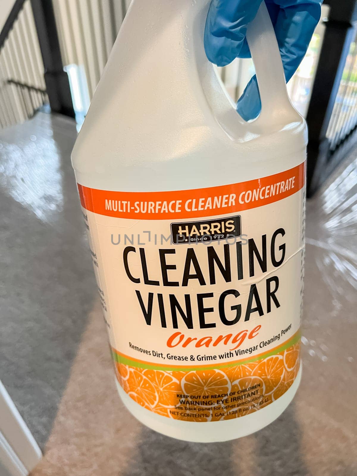 Castle Rock, Colorado, USA-July 15, 2023-A person wearing a blue glove holds a large bottle of Harris Cleaning Vinegar Orange multi-surface cleaner. The label indicates it removes dirt, grease, and grime using vinegar cleaning power. The background shows a carpeted floor covered with protective plastic, suggesting a cleaning or home maintenance setting.