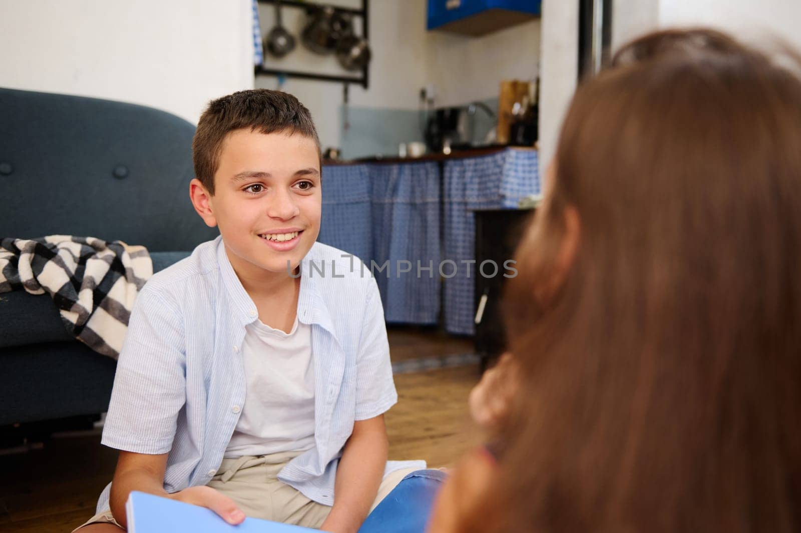 A cheerful boy sitting on the floor in a cozy kitchen, smiling warmly during a conversation with a girl in the foreground.