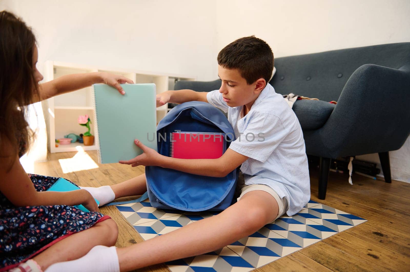 Two children prepare for school by packing notebooks into a school bag, sitting indoors.