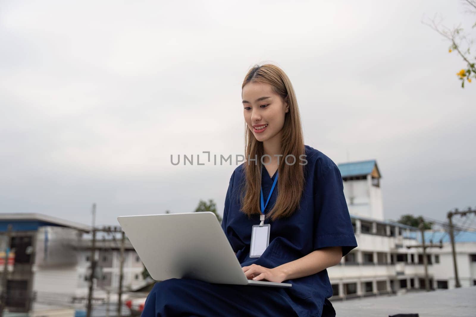 A cheerful female healthcare professional in scrubs smiles while using a laptop outdoors, emphasizing the importance of relaxation and modern technology in her role.