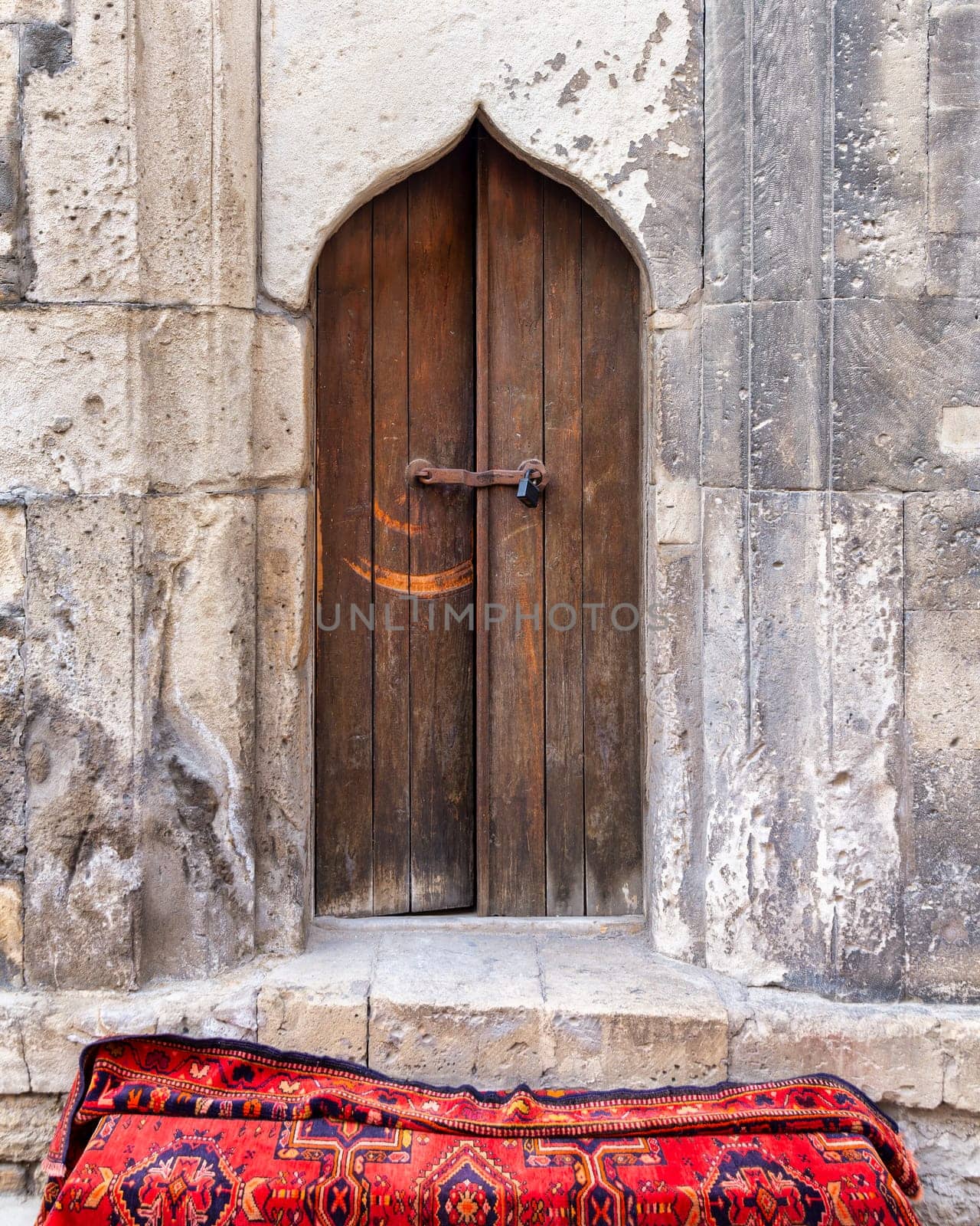 An ornate doorway in a textured stone wall with intricate carvings, a vibrant red rug on cobblestones, highlight the charm of old Baku, Azerbaijan, during the first National Carpet Festival