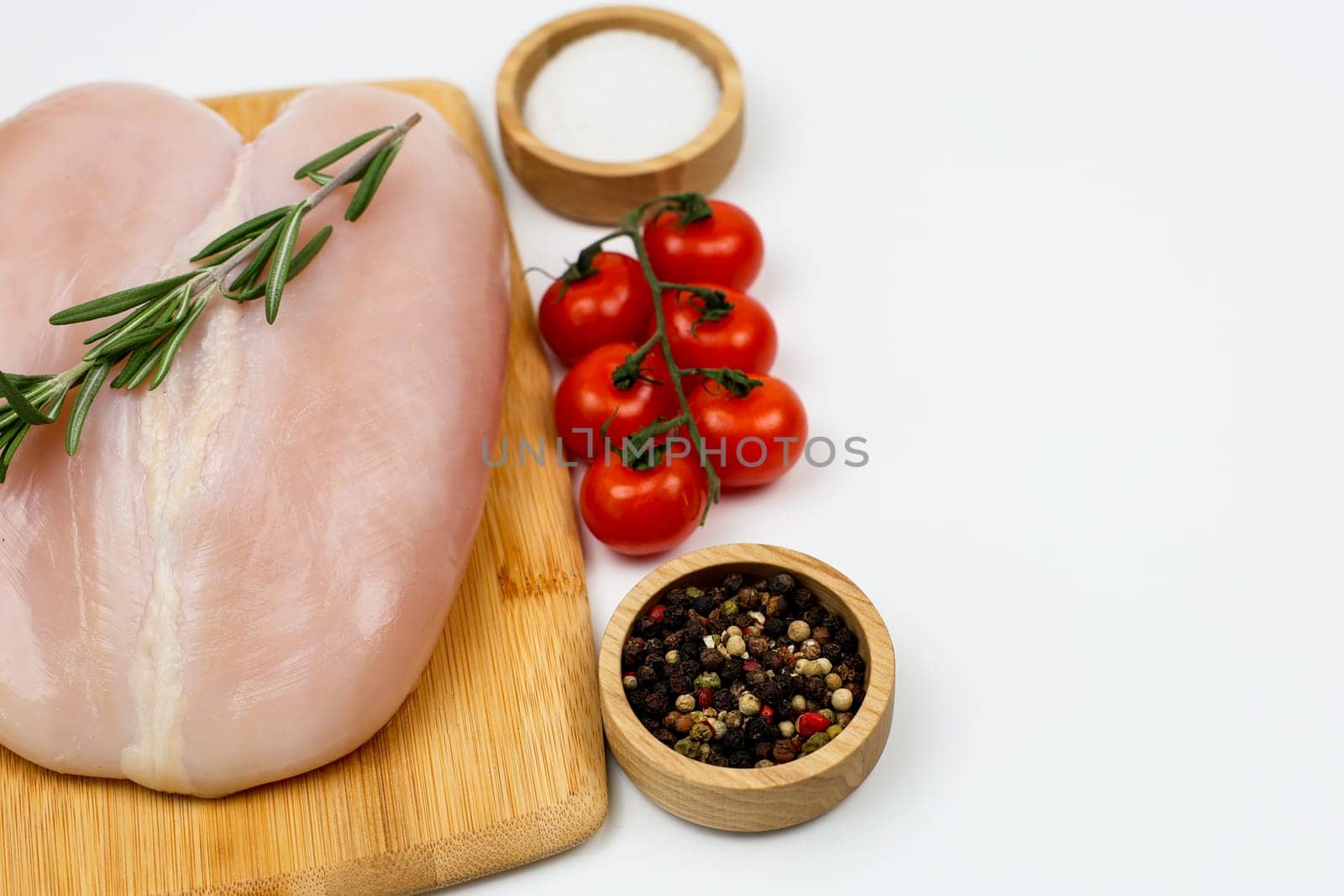 A close-up on the left of a chicken fillet with a sprig of rosemary lies on a wooden cutting board, next to spices and salt in wooden bowls and a sprig of tomatoes on a white background with space for text. The concept of cooking, dietary menu, healthy and natural products. High quality photo