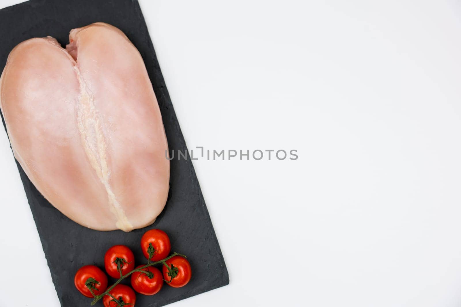 Close-up view from top left: chicken fillet with a sprig of tomatoes next to it lies on a black stone board on a white background with space for text. The concept of cooking, dietary menu, healthy and natural products. High quality photo