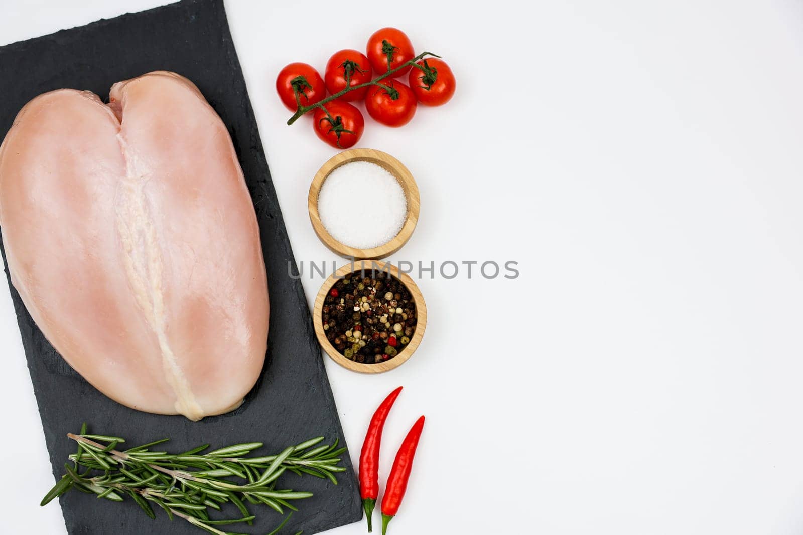 Top view of chicken fillet on a black stone board with a sprig of rosemary, next to a sprig of tomatoes, spices and salt in wooden bowls and red hot peppers on a white background with space for text. The concept of cooking, dietary menu, healthy and natural products. High quality photo