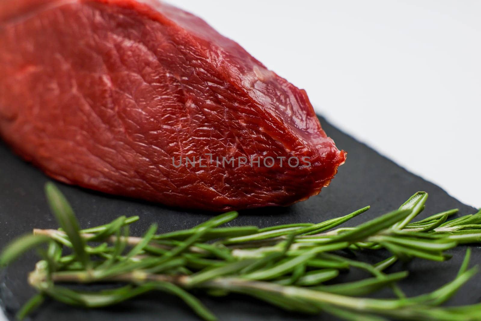 Close-up of red beef tenderloin on black stone plank with a sprig of rosemary on a white background. The concept of cooking, dietary menu, healthy and natural products. High quality photo